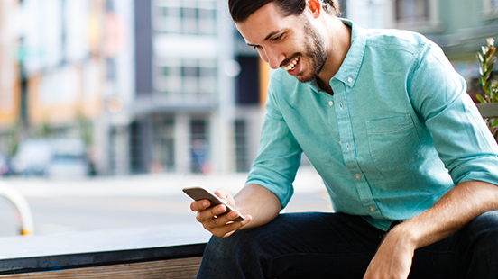 Man seated outside on city street using mobile phone.