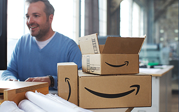 A smiling business owner on a computer surrounded by multiple Amazon cardboard shipping boxes.