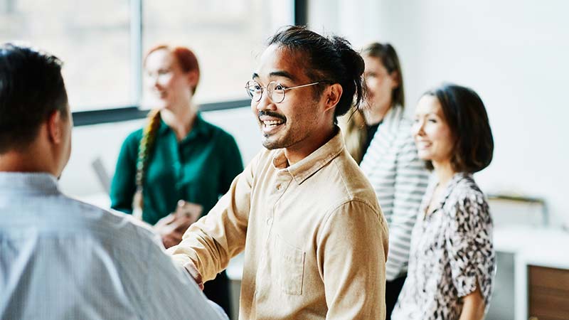 Office employee shakes another employee's hand in a meeting while others converse in the background.